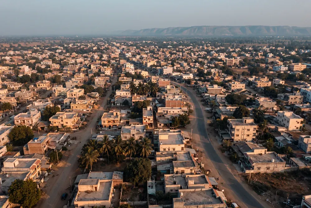 A North Gujarat town seen from the air at golden hour, low flat-roofed buildings in warm ochre with palm trees and a road curving through.