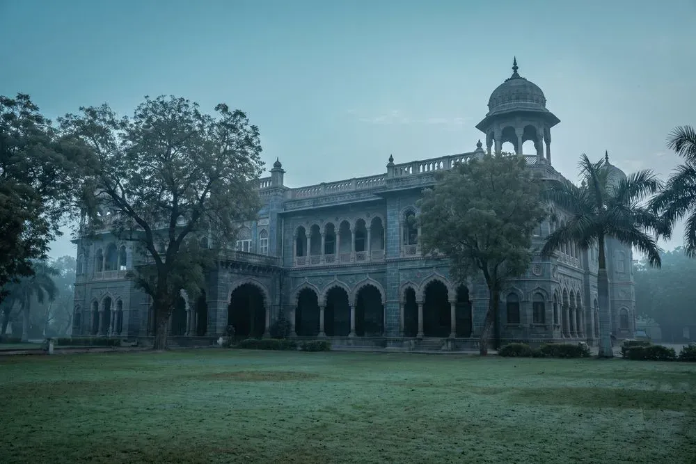 A nineteenth-century Indo-Saracenic museum building in Rajkot at dawn, its arched stone arcade and domed corner pavilion standing behind the trees and lawns of an old public garden.
