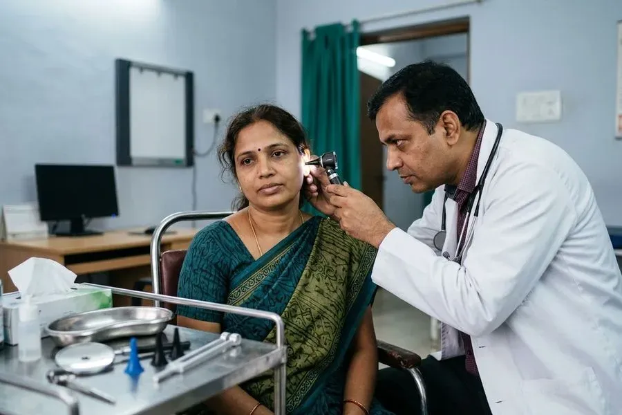 An ENT doctor examining the ear of a seated woman patient with a handheld otoscope in a tidy consulting room.